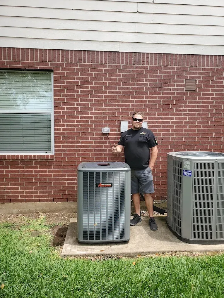 Technician standing beside two air conditioning units against a brick wall.